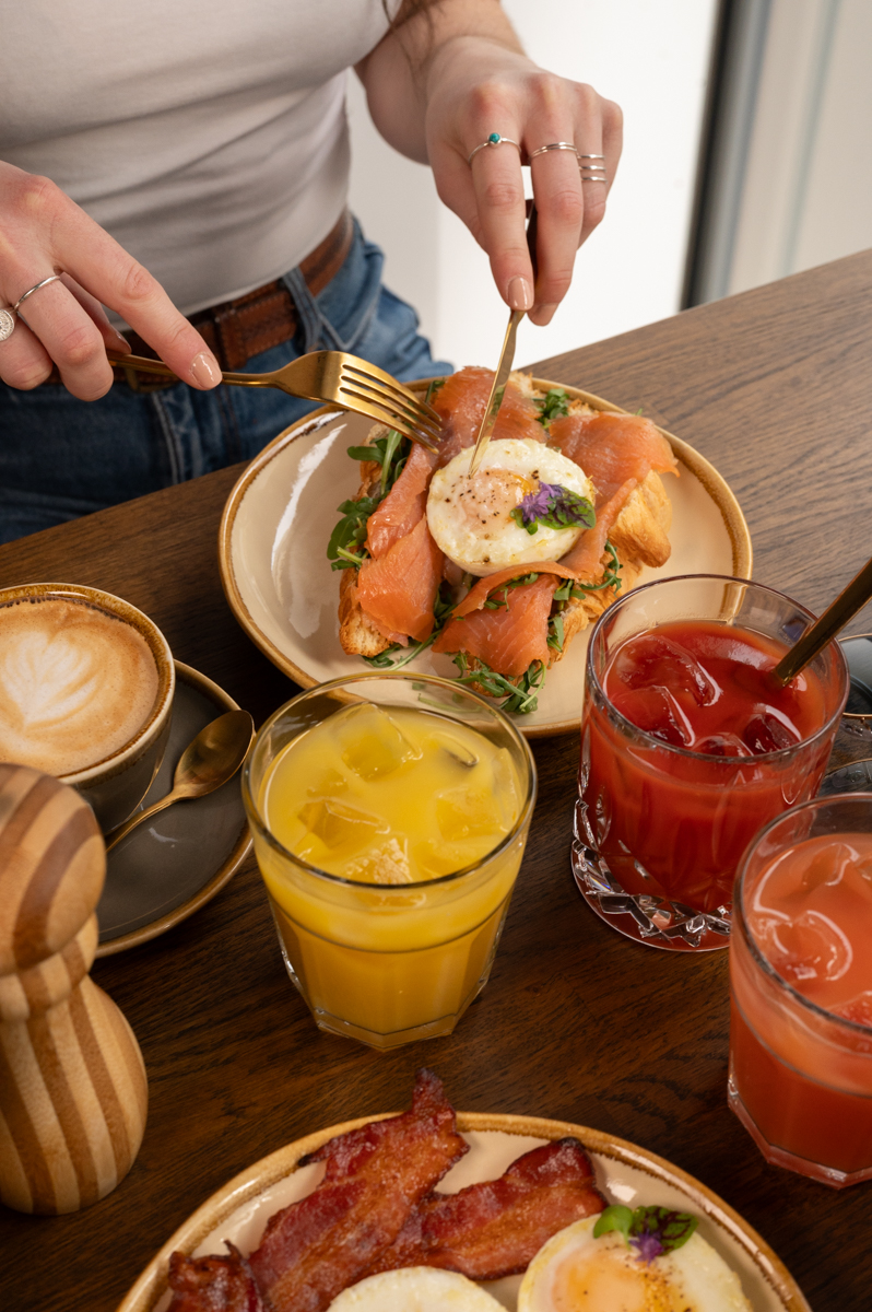Gast isst ein Croissant mit Lachs und Ei, umgeben von Kaffee, Säften und Speck